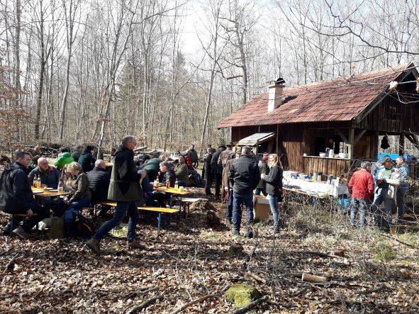 Viele Menschen sitzen neben einer Hütte im Wald auf Bierbänken