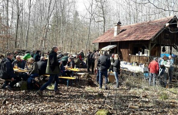 Viele Menschen sitzen neben einer Hütte im Wald auf Bierbänken