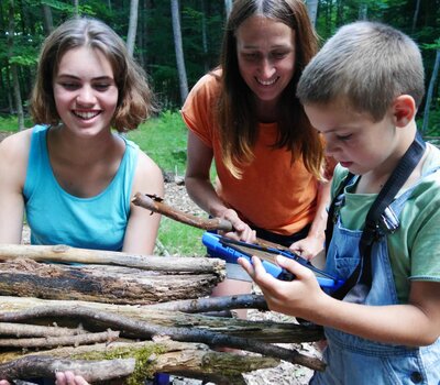 Zwei Kinder und eine Betreuerin im Wald mit Stöcken